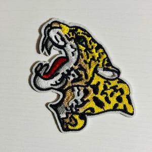 May include: An embroidered iron-on patch featuring a yellow, black, and white jaguar with a red mouth, green eyes, and a white background.