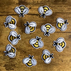 May include: Twelve white and yellow embroidered bee patches. The bees have black eyes and a yellow and black striped abdomen.