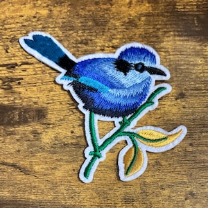 May include: Embroidered patch featuring a blue bird perched on a green branch with yellow leaves. The bird has a blue body, black beak, and a white border. The patch is designed for application on clothing or accessories.
