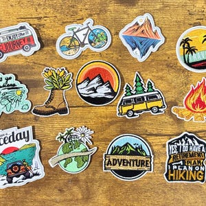 May include: A collection of 11 embroidered iron-on patches featuring various travel and adventure themes. The patches include images of a camper van, a bicycle, a mountain range, a campfire, a globe, and a globe with the text "Let's go somewhere".