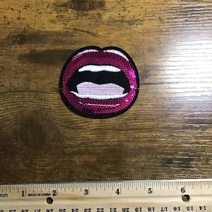 May include: A sequined patch in the shape of open lips, with a dark pink color and a black outline. The lips are open, revealing white teeth and a black interior. A ruler is placed below the patch.