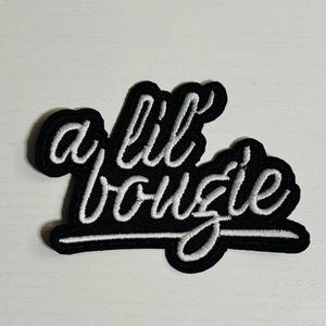 May include: Black patch with white embroidered text that reads "a lil bougie". The patch has a black border and is rectangular in shape. The text is in a cursive font.