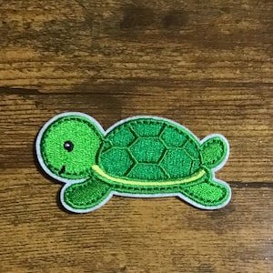May include: Green embroidered turtle patch with a white outline. The turtle has a shell with a hexagonal pattern and a smiling face.