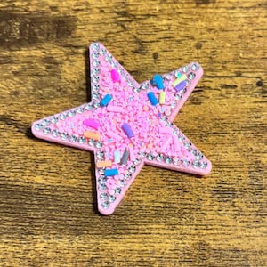 May include: Pink star-shaped embellishment with a sprinkle design and a rhinestone border. The star is covered in small pink sprinkles and accented with colorful sprinkles. The rhinestones are clear and add a touch of sparkle.
