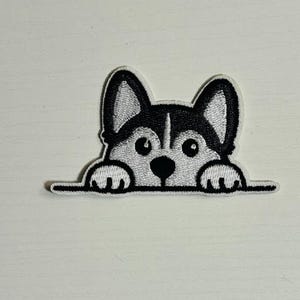 May include: Embroidered patch featuring a cartoon husky dog peeking over a line. The dog has black and white markings, with large eyes and paws visible. The patch is designed for application on clothing or accessories.