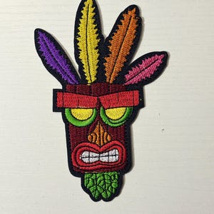 May include: Embroidered patch featuring a stylized tribal mask with a red, brown, and green color scheme. The mask has a wide grin, green eyes, and colorful feathers in purple, yellow, orange, and pink.