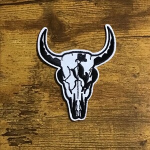 May include: Black and white embroidered patch of a bull skull with cracked details. The patch is on a wooden surface.