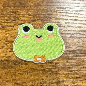 May include: Embroidered patch featuring a smiling green frog with black eyes, pink cheeks, and a small orange bow tie. The frog has a white border and is set against a wooden background. This patch is perfect for adding to clothing or accessories.