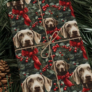 May include: Wrapped gift with festive wrapping paper featuring Weimaraner dogs wearing red scarves, surrounded by greenery and red berries. The gift is tied with twine and set against a backdrop of pine needles and pine cones.