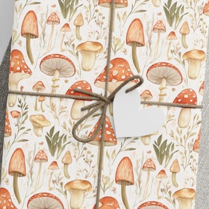 Puede incluir: Papel de regalo blanco con un patrón repetido de setas de acuarela en tonos rojos, naranjas y marrones. Las setas son de varios tamaños y formas, y algunas tienen manchas blancas. El papel está atado con un lazo de cuerda marrón y una etiqueta blanca en forma de corazón.