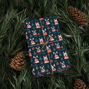 May include: A wrapped gift featuring a repeating pattern of French bulldogs wearing red scarves, Christmas trees, and ornaments on a dark blue background. The gift is tied with twine and set against a backdrop of pine needles and pine cones.