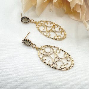 May include: A pair of gold-coloured filigree earrings. Each earring has a small, oval-shaped, grey gemstone stud connected to a larger, openwork oval pendant with a swirling design. The earrings are displayed on a white surface with a soft, floral background.