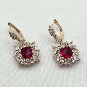 Puede incluir: Pendientes de aro dorados con una piedra cuadrada roja rodeada de piedras transparentes más pequeñas.