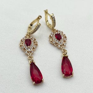 May include: A pair of gold dangle earrings with red teardrop-shaped stones and clear cubic zirconia accents. The earrings feature a delicate filigree design.