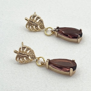 Könnte beinhalten: Ein Paar goldfarbene Ohrringe mit einem Blattdesign und einem tropfenförmigen violetten Edelstein.