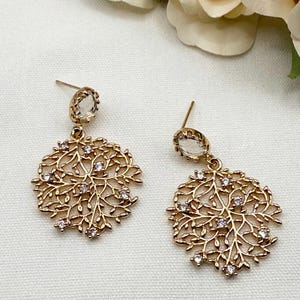 Peut inclure: Une paire de boucles d'oreilles dorées. Chaque boucle d'oreille présente une pierre transparente de forme ovale en haut, reliée à un motif circulaire en filigrane avec des pierres transparentes. Le motif en filigrane ressemble à un motif de branches d'arbres délicates.