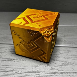 May include: A gold cube-shaped box with intricate geometric patterns and a raised eye symbol on one side.