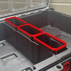 May include: Black storage container with three rectangular compartments edged in red. The container is open, showing a clear lid and a textured interior. The compartments are for organising tools or supplies.