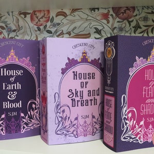 May include: Three hardcover books from the "Crescent City" series, featuring titles "House of Earth & Blood", "House of Sky and Breath", and "House of Flame and Shadow". Each book has a unique color scheme with ornate designs and text.