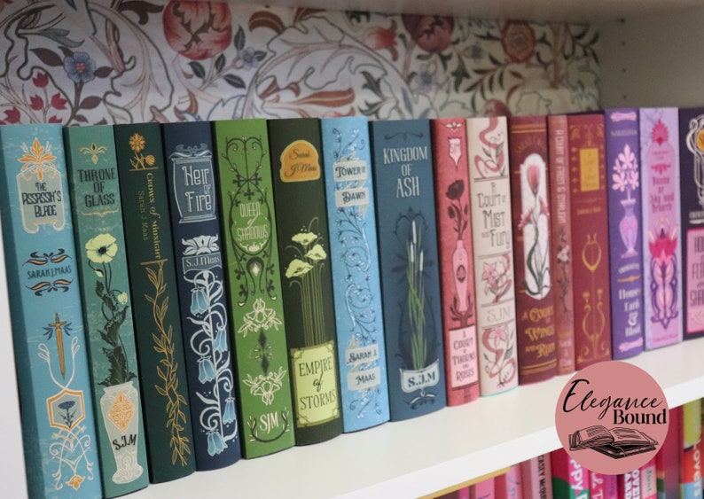 Pode incluir: Uma cole&ccedil;&atilde;o de livros decorativos com lombadas coloridas, dispostos numa prateleira branca. Os livros apresentam desenhos florais e geom&eacute;tricos, com t&iacute;tulos como "Throne of Glass" e "Heir of Fire". O fundo &eacute; um papel de parede floral.