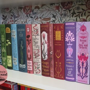 May include: A collection of fantasy novels with decorative covers, arranged on a white shelf. The books feature various colors, including blue, green, red, and purple, with floral and geometric designs. The text "Elegance Bound" is in a pink circle.
