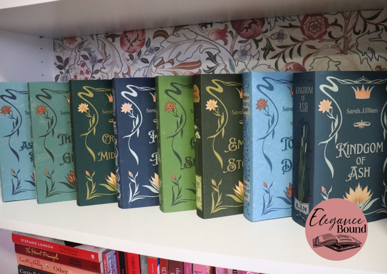 Pode incluir: Uma cole&ccedil;&atilde;o de livros de capa dura com desenhos florais e t&iacute;tulos como "Kingdom of Ash" e "Throne of Glass" numa prateleira branca. As lombadas s&atilde;o em v&aacute;rios tons de verde e azul, com detalhes dourados. O fundo apresenta um papel de parede floral.