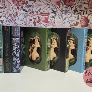 Pode incluir: Uma cole&ccedil;&atilde;o de livros de capa dura com capas decorativas. As lombadas apresentam t&iacute;tulos e nomes de autores em v&aacute;rias fontes. As capas apresentam desenhos florais e um retrato de perfil de uma mulher em estilo art nouveau. Os livros est&atilde;o dispostos numa superf&iacute;cie branca.