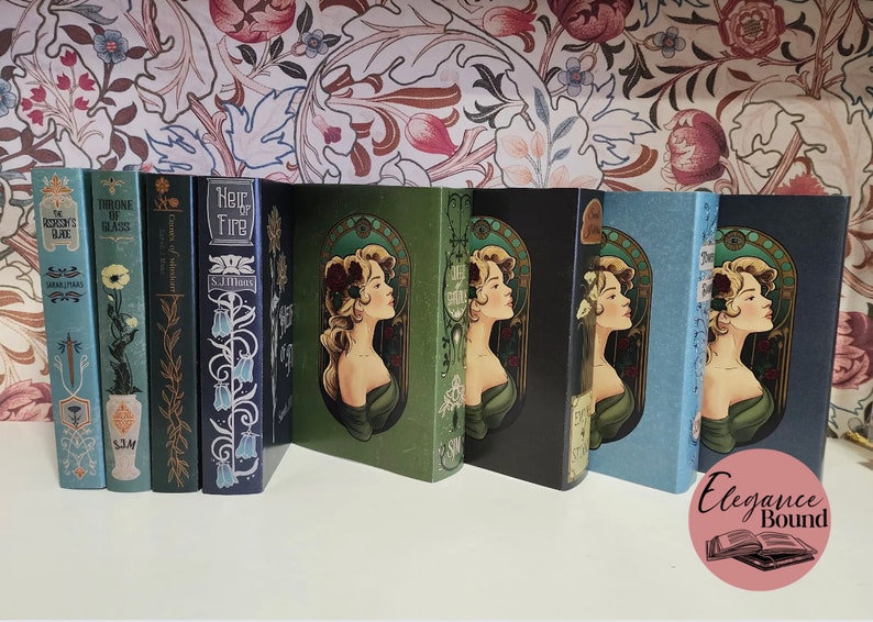 Pode incluir: Uma cole&ccedil;&atilde;o de livros de capa dura com capas decorativas. As lombadas apresentam t&iacute;tulos e nomes de autores em v&aacute;rias fontes. As capas apresentam desenhos florais e um retrato de perfil de uma mulher em estilo art nouveau. Os livros est&atilde;o dispostos numa superf&iacute;cie branca.