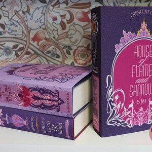 May include: A stack of three hardcover books with decorative covers. The books are in shades of purple and pink, with the titles "House of Flame and Shadow", "House of Sky and Breath", and "House of Earth and Blood" visible. The spines feature the text "Crescent City".