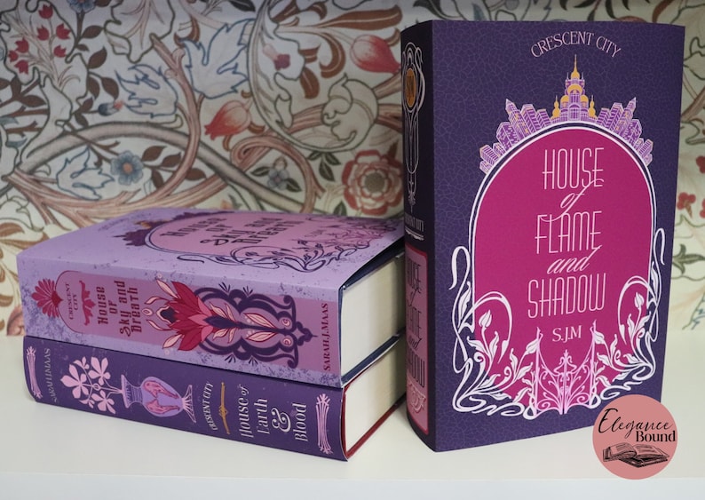 May include: A stack of three hardcover books with decorative covers. The books are in shades of purple and pink, with the titles "House of Flame and Shadow", "House of Sky and Breath", and "House of Earth and Blood" visible. The spines feature the text "Crescent City".