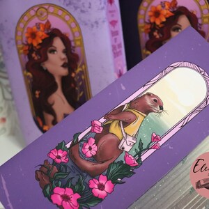May include: Purple bookmark featuring an illustration of an otter wearing a yellow shirt and a white bag, surrounded by pink flowers. The bookmark also features a woman with flowers in her hair. The Elegance Bound logo is visible.