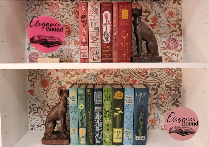 Pode incluir: Uma estante exibe uma cole&ccedil;&atilde;o de livros decorativos com desenhos florais e bot&acirc;nicos. Os livros s&atilde;o em v&aacute;rias cores, incluindo vermelho, verde e azul. Duas est&aacute;tuas de c&atilde;es de bronze s&atilde;o colocadas nas prateleiras. O texto "Elegance Bound" est&aacute; em um c&iacute;rculo rosa.