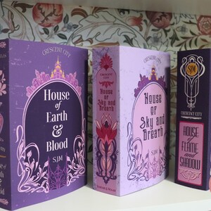 May include: Three hardcover books with matching designs. The books are titled "House of Earth & Blood", "House of Sky and Breath", and "House of Flame and Shadow". Each book has a purple cover with ornate floral and architectural details.