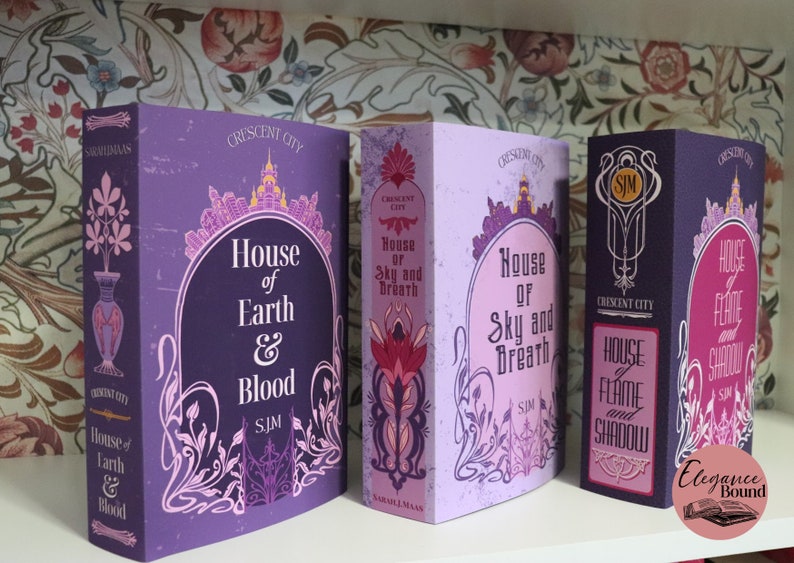 May include: Three hardcover books with matching designs. The books are titled "House of Earth & Blood", "House of Sky and Breath", and "House of Flame and Shadow". Each book has a purple cover with ornate floral and architectural details.