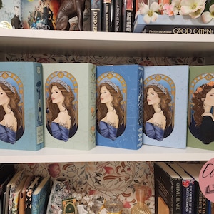 May include: A collection of hardcover books with illustrated covers featuring a woman with long brown hair and a crown. The books are in various shades of blue and green, arranged on a white bookshelf.