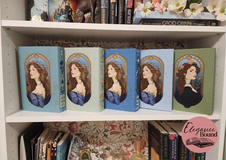 May include: A collection of hardcover books with illustrated covers featuring a woman with long brown hair and a crown. The books are in various shades of blue and green, arranged on a white bookshelf.