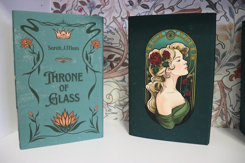 Pode incluir: Dois livros de capa dura. O livro da esquerda &eacute; verde-azulado com o t&iacute;tulo "Throne of Glass" e o nome do autor. O livro da direita &eacute; verde escuro com um retrato de mulher e rosas vermelhas. Ambos os livros t&ecirc;m elementos florais decorativos.