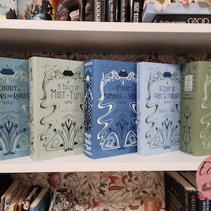 May include: Five hardcover books with matching covers are displayed on a white shelf. The books are in shades of blue and green, with decorative floral and mountain designs. The titles include "A Court of Thorns and Roses" and "A Court of Mist and Fury."
