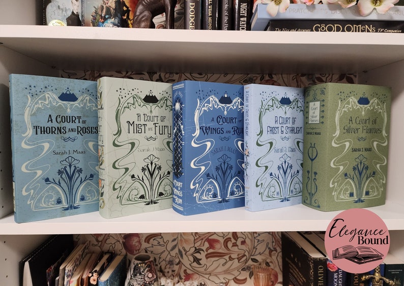 May include: Five hardcover books with matching covers are displayed on a white shelf. The books are in shades of blue and green, with decorative floral and mountain designs. The titles include "A Court of Thorns and Roses" and "A Court of Mist and Fury."