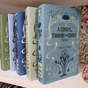 May include: A row of hardcover books with decorative covers. The books are in shades of blue and green, with floral and abstract designs. The title "A Court of Thorns and Roses" is visible on one book.