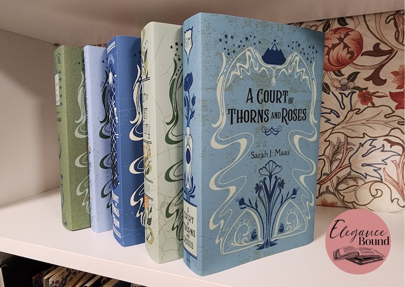 May include: A row of hardcover books with decorative covers. The books are in shades of blue and green, with floral and abstract designs. The title "A Court of Thorns and Roses" is visible on one book.