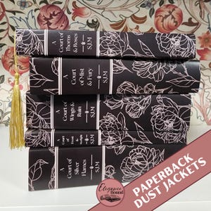 May include: A stack of five black paperback books with white floral designs. The spines display titles like "A Court of Thorns & Roses" and "A Court of Mist & Fury." A gold tassel hangs from the top book. The text "PAPERBACK DUST JACKETS" is visible.
