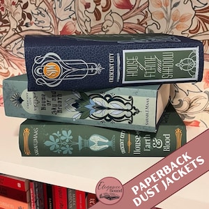 Pode incluir: Uma pilha de três livros de capa dura com sobrecapas decorativas. Os livros são em tons de azul e verde, com detalhes dourados e brancos. Os títulos incluem "House of Flame and Shadow", "House of Sky and Breath" e "House of Earth and Blood."