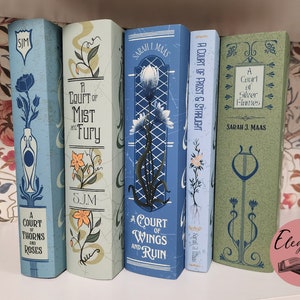 May include: A collection of fantasy novels with decorative covers. The books are in shades of blue and green, with floral and art nouveau-inspired designs. Titles include "A Court of Thorns and Roses" and "A Court of Wings and Ruin."