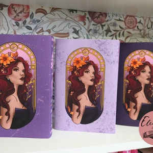May include: Three purple journals with a woman illustration. The woman has red hair and wears a black dress, adorned with a floral crown. The journals are displayed on a white shelf against a floral wallpaper background. The journals are labeled "Elegance Bound".