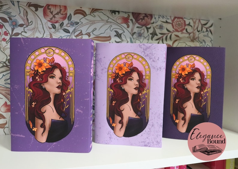 May include: Three purple journals with a woman illustration. The woman has red hair and wears a black dress, adorned with a floral crown. The journals are displayed on a white shelf against a floral wallpaper background. The journals are labeled "Elegance Bound".