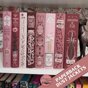Könnte beinhalten: Eine Sammlung von Taschenbuch-Schutzumschlägen in Rosa- und Burgundertönen, angeordnet auf einem weißen Regal. Die Umschläge weisen aufwendige Blumen- und Zierdesigns mit Titeln und Autorennamen auf. Der Text "PAPERBACK DUST JACKETS" ist im unteren rechten Eck sichtbar.