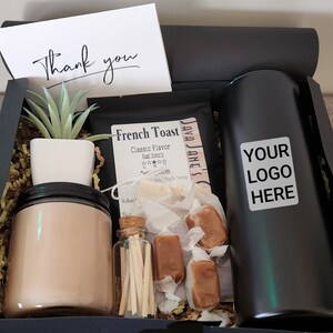 Puede incluir: Una caja de regalo negra llena de varios artículos, incluyendo una vela, cerillas, caramelos de caramelo, una pequeña planta en maceta, una bolsa de mezcla para tostadas francesas y una taza de viaje negra con el texto "YOUR LOGO HERE".