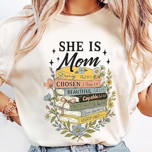 May include: White t-shirt with a graphic of a stack of books with floral accents and the text "She is Mom" with bible verses on each book. The text on the books reads: "Strong Phil 4:13", "Chosen 1 Thess 1:4", "Beautiful Tell 3:11", "Capable Mark 10:27", "Victorious Rom 8:13", "Never alone Mat 28:20", "Greatly Loved Jon 3:16"