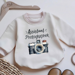 May include: Cream-colored baby sweatshirt with the words "Assistant Photographer" and a vintage camera graphic. The sweatshirt has a light pink trim around the neck and cuffs. A wooden hanger with the word "libellulas" is visible.